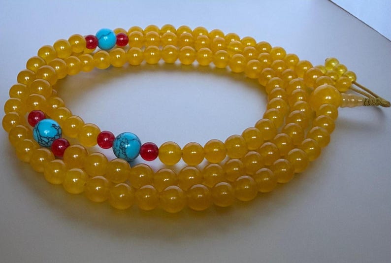 May include: A long strand of prayer beads featuring smooth, translucent yellow beads. The strand is accented with three larger, round, bright turquoise beads with dark veining, each flanked by two smaller, vibrant red spherical beads.