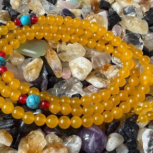 May include: A long strand of round yellow beads featuring interspersed bright turquoise stone beads and small red accent beads. The strand is finished with a larger yellow bead and a yellow cord tassel at one end, creating a continuous loop.