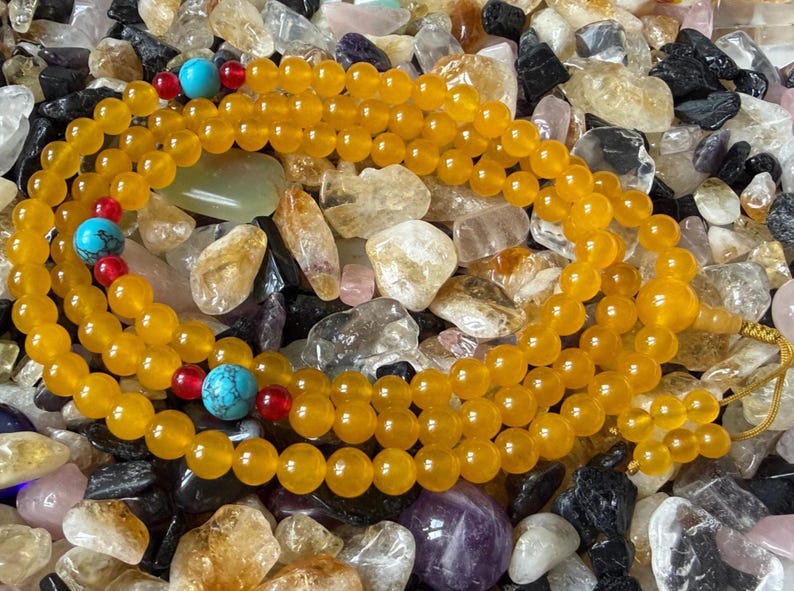 May include: A long strand of round yellow beads featuring interspersed bright turquoise stone beads and small red accent beads. The strand is finished with a larger yellow bead and a yellow cord tassel at one end, creating a continuous loop.