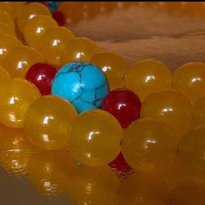 May include: A multi-strand necklace featuring smooth, round yellow beads interspersed with smaller, glossy red beads and a single, prominent turquoise-colored bead with dark veining. The beads are arranged in a repeating pattern on a string.