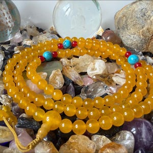 May include: A long strand of translucent yellow prayer beads featuring round polished stones. The necklace is accented with small red beads and larger turquoise-colored beads at intervals, finished with a yellow cord tassel at the end.