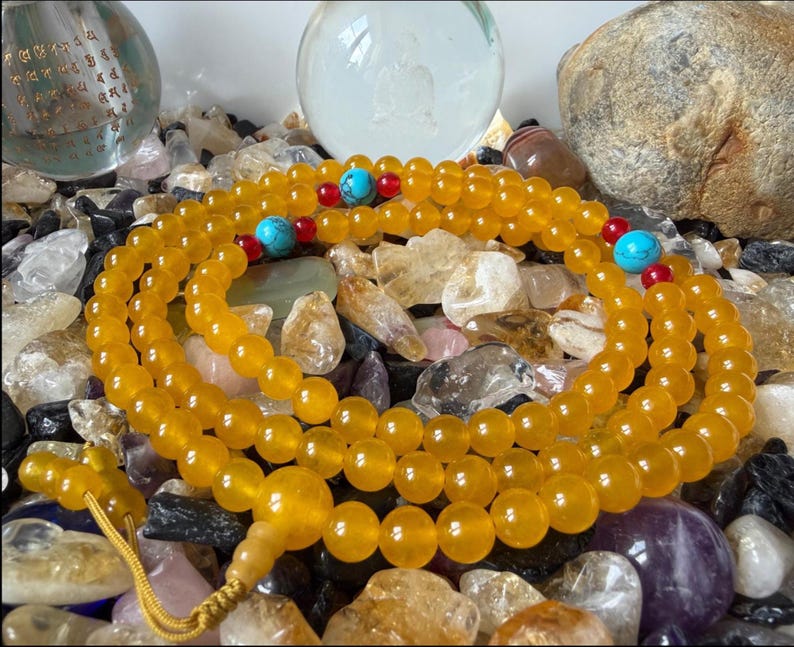 May include: A long strand of translucent yellow prayer beads featuring round polished stones. The necklace is accented with small red beads and larger turquoise-colored beads at intervals, finished with a yellow cord tassel at the end.