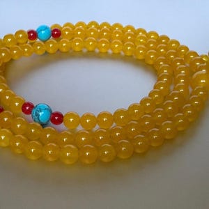 May include: A long strand of yellow beads arranged in a circular pattern. The strand features interspersed accents of bright turquoise blue spherical beads and smaller red spherical beads, with a knotted cord end and a larger yellow bead.