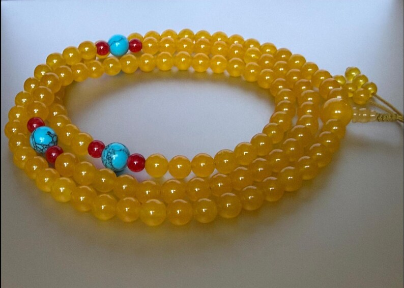 May include: A long strand of yellow beads arranged in a circular pattern. The strand features interspersed accents of bright turquoise blue spherical beads and smaller red spherical beads, with a knotted cord end and a larger yellow bead.