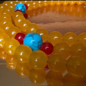 May include: A beaded necklace featuring smooth, round, translucent yellow beads arranged in a circular pattern. The strand is accented with vibrant turquoise blue beads with dark veining and small, glossy, solid red spherical beads.