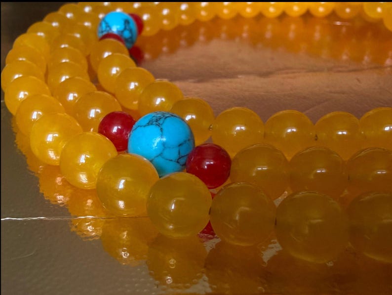 May include: A beaded necklace featuring smooth, round, translucent yellow beads arranged in a circular pattern. The strand is accented with vibrant turquoise blue beads with dark veining and small, glossy, solid red spherical beads.