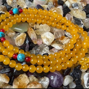 May include: A prayer bead necklace featuring smooth, round, translucent yellow beads arranged in a continuous loop. The strand is accented with three larger, round, bright blue turquoise beads, each flanked by two small, vibrant red spherical beads.