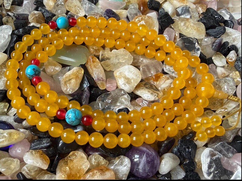 May include: A prayer bead necklace featuring smooth, round, translucent yellow beads arranged in a continuous loop. The strand is accented with three larger, round, bright blue turquoise beads, each flanked by two small, vibrant red spherical beads.