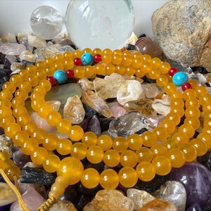 May include: A strand of translucent yellow beads arranged in a circular pattern, featuring small red accent beads and larger round turquoise-colored beads interspersed throughout the length, finished with a yellow cord and a larger yellow bead.