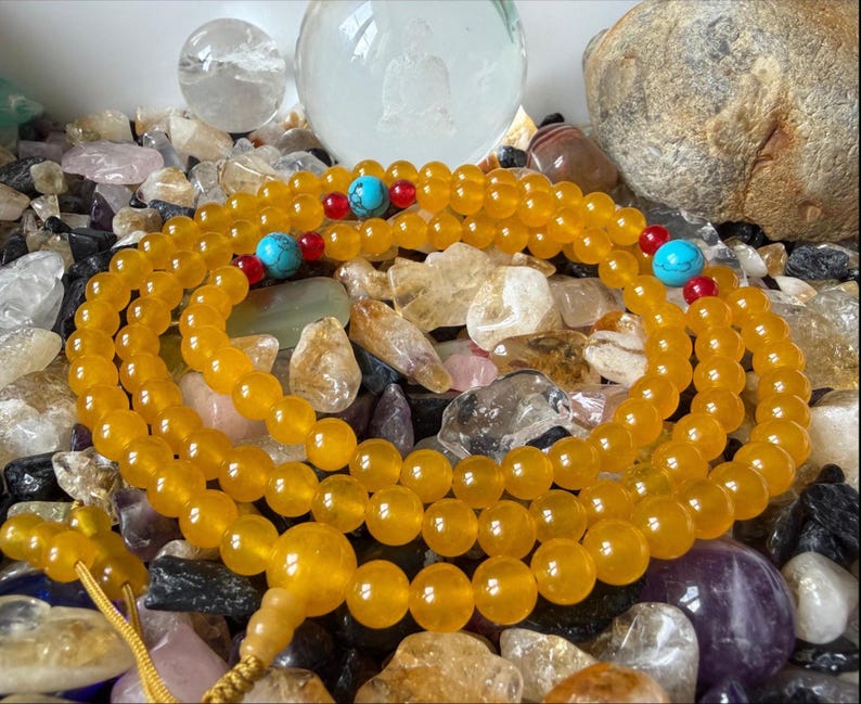 May include: A strand of translucent yellow beads arranged in a circular pattern, featuring small red accent beads and larger round turquoise-colored beads interspersed throughout the length, finished with a yellow cord and a larger yellow bead.