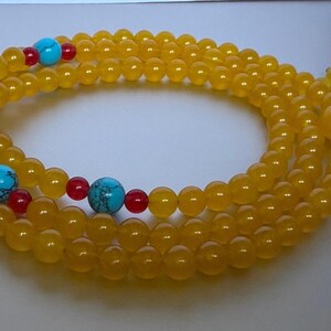 May include: A long strand of translucent yellow beads arranged in a circular pattern. The necklace features interspersed accents of bright turquoise blue spherical beads with dark veining, flanked by smaller, smooth, opaque red spherical beads.
