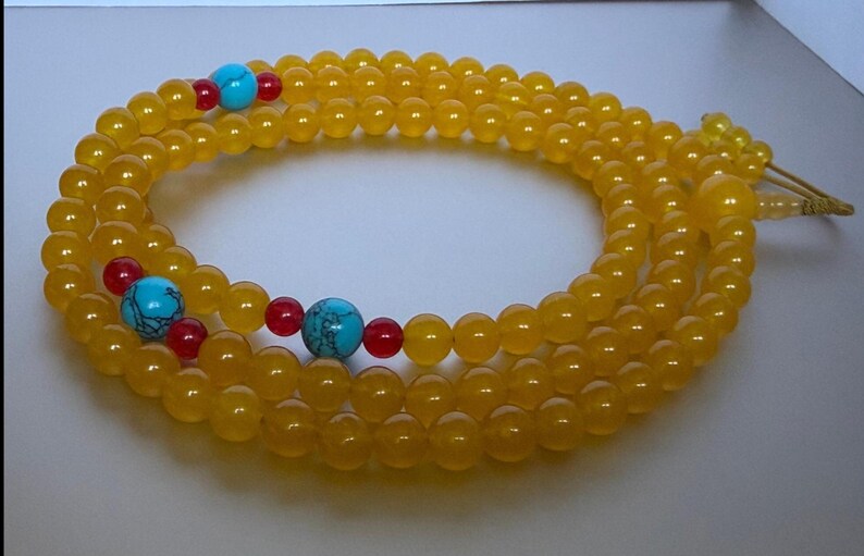 May include: A long strand of translucent yellow beads arranged in a circular pattern. The necklace features interspersed accents of bright turquoise blue spherical beads with dark veining, flanked by smaller, smooth, opaque red spherical beads.