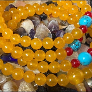 May include: A beaded necklace featuring translucent yellow spherical beads arranged in multiple loops. The strand is accented with two larger bright turquoise blue beads and four smaller deep red beads interspersed along the yellow bead chain.