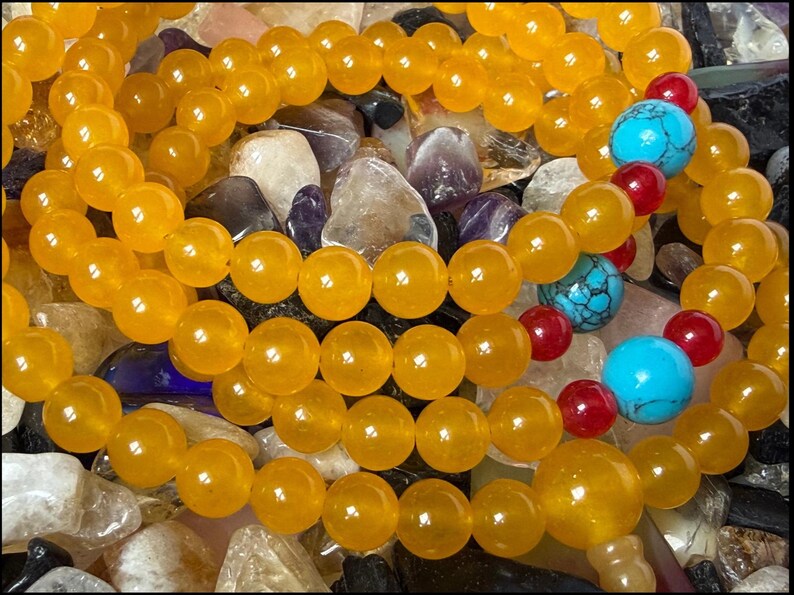 May include: A beaded necklace featuring translucent yellow spherical beads arranged in multiple loops. The strand is accented with two larger bright turquoise blue beads and four smaller deep red beads interspersed along the yellow bead chain.