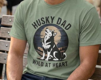 May include: A light green t-shirt with the text "HUSKY DAD WILD AT HEART" and a graphic of a husky howling at the moon. The shirt is being worn by a person sitting on a park bench. Several dogs are in the background.