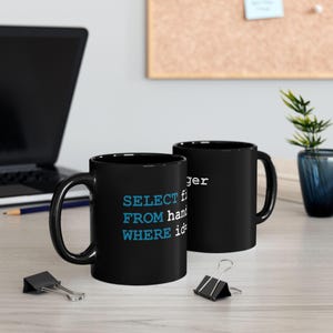 May include: Two black coffee mugs with white and blue text. One mug displays "SELECT FROM hand WHERE id" in blue, the other displays "ger" in white. The mugs are on a light-coloured surface.