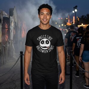 May include: Black t-shirt with a white graphic of a smiling face with stitched eyes and the text "Smells Like Halloween Spirit". The shirt is worn by a person in an outdoor setting with other people and structures in the background.