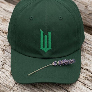 May include: A dark forest green baseball cap featuring a centred, embroidered bright green letter W in a gothic-style font. A single sprig of dried purple lavender is placed horizontally across the centre of the cap's curved brim.