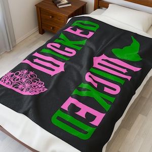 Peut inclure: Couverture noire avec le mot "WICKED" en rose et vert, ainsi qu'une tiare rose et un chapeau de sorcière vert. La couverture est posée sur un lit avec un oreiller blanc et une table de chevet en bois.