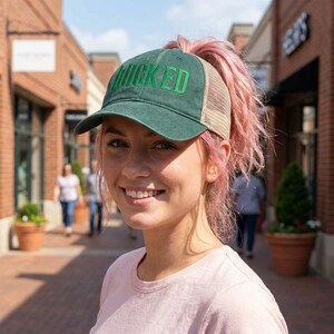 May include: A green baseball cap with the word "WICKED" embroidered in green. The cap has a mesh back and a curved brim. The person in the image is wearing a pink shirt.