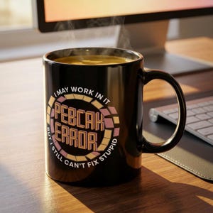 May include: Black ceramic mug with a curved handle, filled with a hot drink. The mug features the text "I MAY WORK IN IT, BUT I STILL CAN'T FIX STUPID" in a circular design with pink and yellow accents.