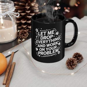 May include: A black ceramic coffee mug with the text "LET ME DROP EVERYTHING AND WORK ON YOUR PROBLEM" in white. The mug has a curved handle and is filled with a dark liquid, possibly coffee or tea. Steam rises from the mug.