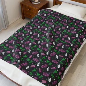 Peut inclure: Couverture noire avec un motif répété d'éléments graphiques blancs, verts et roses. Le motif comprend des chapeaux de sorcière stylisés, des cercueils et d'autres symboles. La couverture est sur un lit avec un drap blanc.