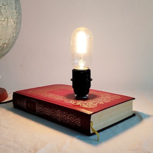 Könnte beinhalten: Eine Glühbirne im Vintage-Stil, die auf einem roten Hardcover-Buch mit goldenen Akzenten montiert ist. Die Glühbirne ist eingeschaltet und beleuchtet das Buch.