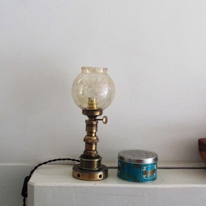 Op de afbeelding: Een messing lamp met een heldere glazen bolvormige kap staat op een wit rek met een blikje kaarsen. De lamp heeft een zwart snoer en een kleine knop aan de zijkant.