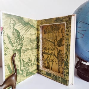 Puede incluir: Un libro abierto con un diseño tridimensional y en capas. Las páginas presentan ilustraciones detalladas en verde y crema de personas en un entorno de jardín. El lomo del libro es blanco y una pequeña figura de dinosaurio de bronce está en primer plano.
