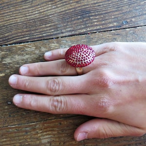 May include: A red ring with a large, round, sparkly red stone. The ring is on a person's finger.