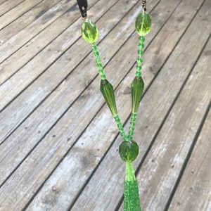 Op de afbeelding: Een groene glazen kralenketting met een kwast. De ketting heeft een zwart lint aan de bovenkant en een grote groene glazen kraal aan de onderkant.