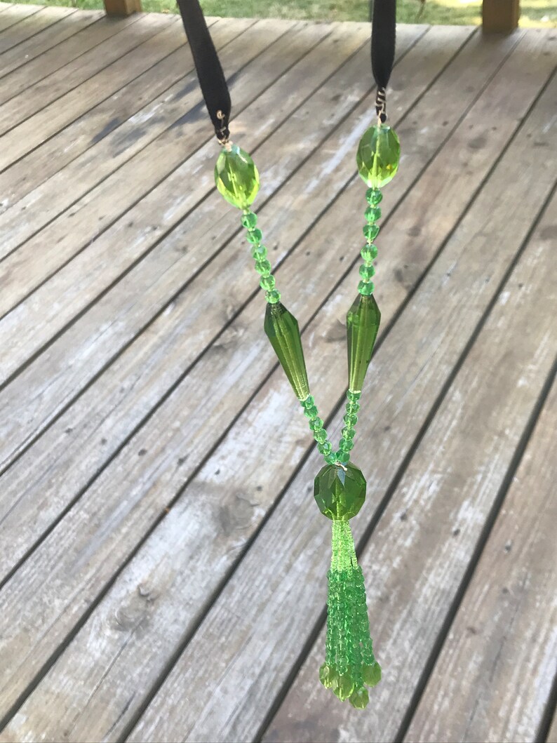 Op de afbeelding: Een groene glazen kralenketting met een kwast. De ketting heeft een zwart lint aan de bovenkant en een grote groene glazen kraal aan de onderkant.