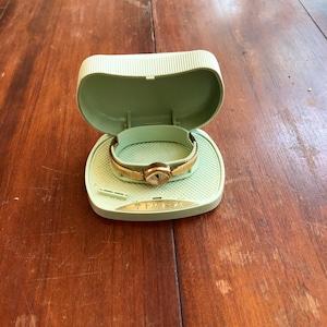 May include: A gold-toned wristwatch with a white face and black hands is displayed in a light green plastic watch box. The box has the word "TIMEX" printed on the bottom.