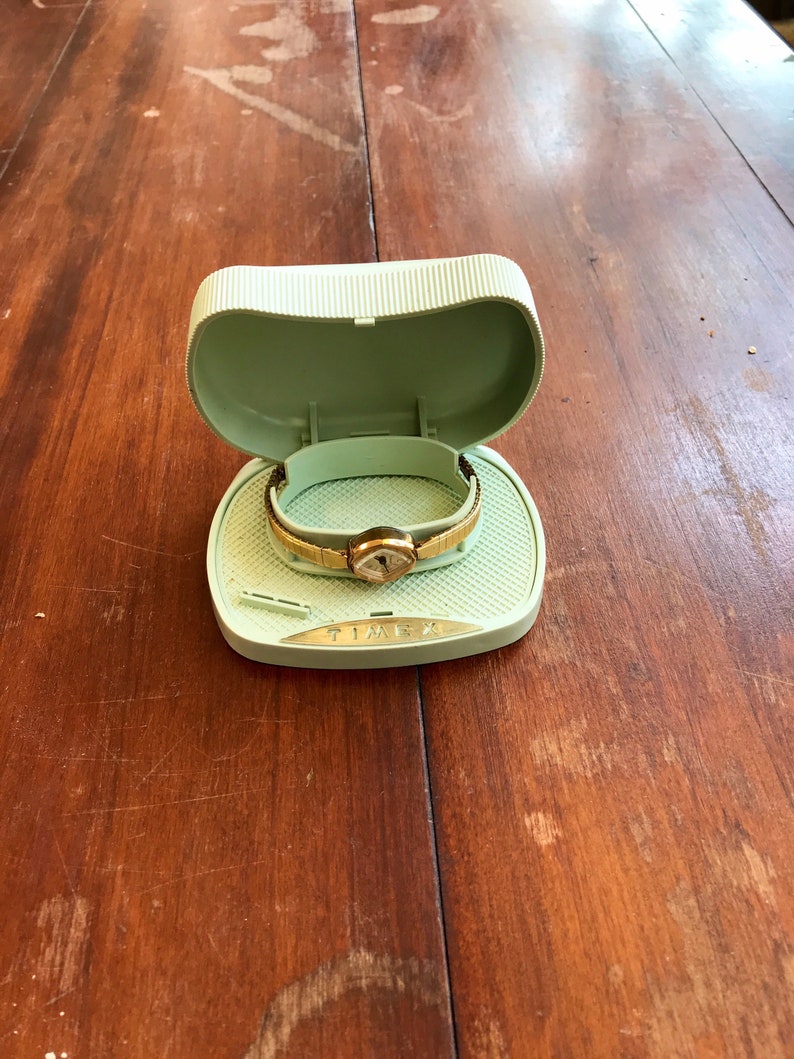May include: A gold-toned wristwatch with a white face and black hands is displayed in a light green plastic watch box. The box has the word "TIMEX" printed on the bottom.