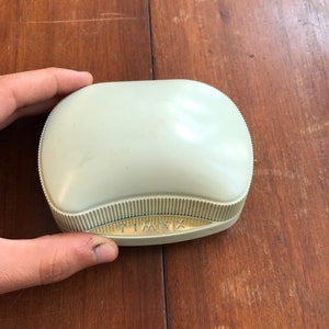 May include: A light green, oval-shaped, plastic box with a gold-colored label that reads "TIMEX".