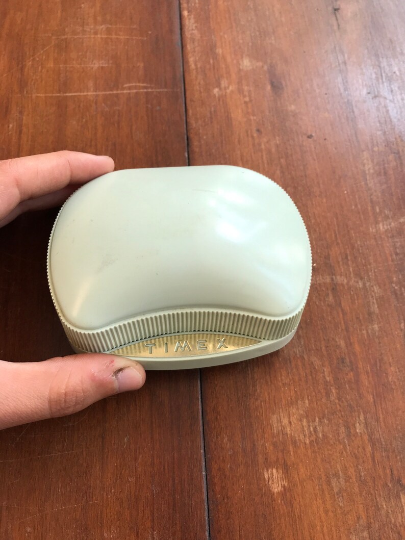 May include: A light green, oval-shaped, plastic box with a gold-colored label that reads "TIMEX".