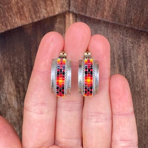 May include: A pair of silver hoop earrings with red, yellow, and black beaded designs. The earrings are made of metal and have a hoop shape.