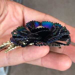Peut inclure: Une broche en forme de fleur noire iris&eacute;e avec un centre en perle. La broche a une &eacute;pingle dor&eacute;e au dos.
