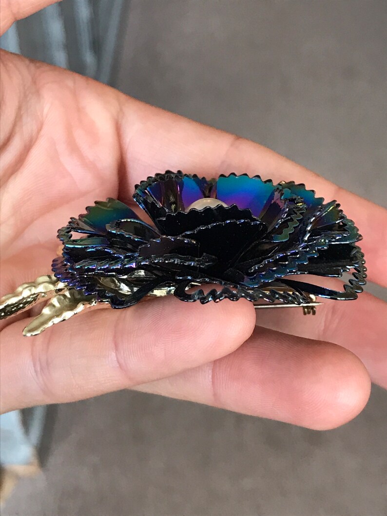 Peut inclure: Une broche en forme de fleur noire iris&eacute;e avec un centre en perle. La broche a une &eacute;pingle dor&eacute;e au dos.