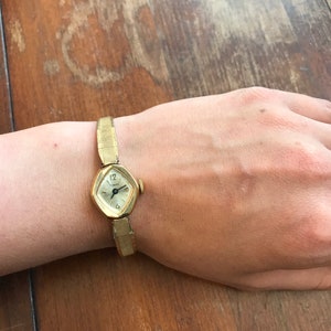 May include: A gold-toned wristwatch with a square face and a metal band. The watch face has black Roman numeral hour markers and black hands.