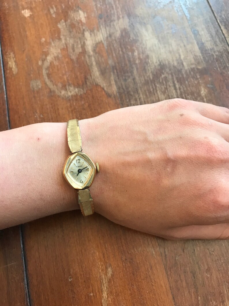 May include: A gold-toned wristwatch with a square face and a metal band. The watch face has black Roman numeral hour markers and black hands.