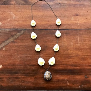 May include: A necklace made of light yellow shell beads with a larger shell pendant in the center. The pendant has a brown and white pattern.