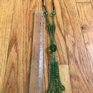 Op de afbeelding: Een lange groene glazen kralenketting met een kwast. De ketting is ongeveer 45 cm lang en heeft een zwarte lintstrik.