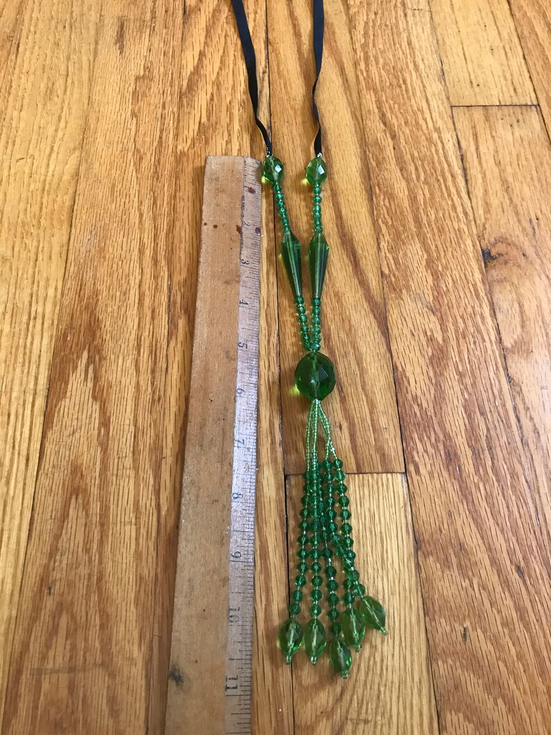 Op de afbeelding: Een lange groene glazen kralenketting met een kwast. De ketting is ongeveer 45 cm lang en heeft een zwarte lintstrik.