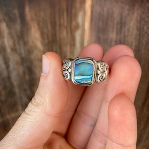 May include: A silver ring featuring a square abalone shell inlay. The ring band is adorned with a swirling design.