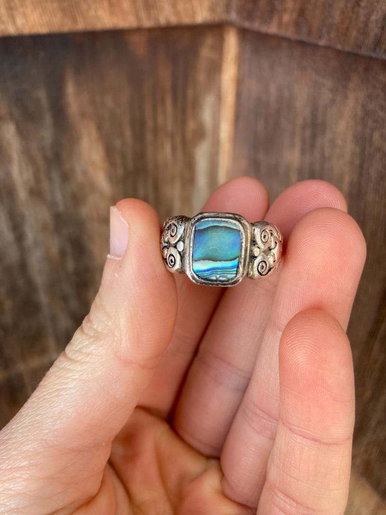 May include: A silver ring featuring a square abalone shell inlay. The ring band is adorned with a swirling design.