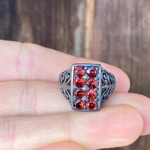May include: A silver ring with a rectangular face set with eight vibrant red gemstones. The ring has an ornate, filigree design on the band and around the gemstone setting. The gemstones are arranged in two rows of four.