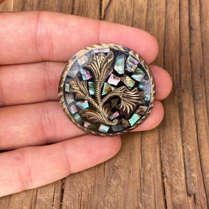 May include: A round, silver-toned brooch with a floral design. The brooch is decorated with iridescent mother-of-pearl pieces.