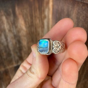 May include: A silver ring featuring a square abalone shell inlay. The ring band has a decorative, swirling design.